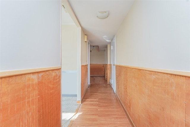 corridor with light wood-type flooring, a wainscoted wall, and wood walls