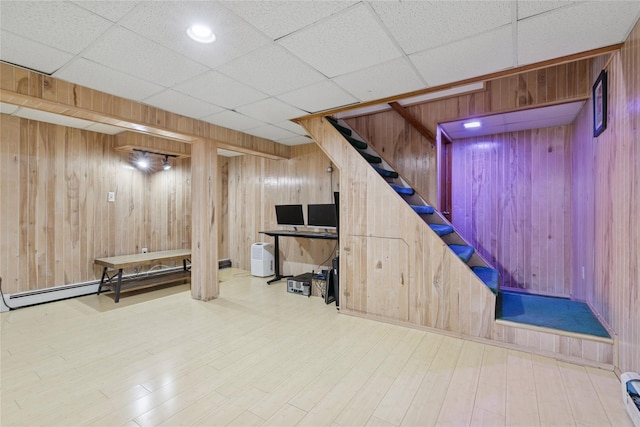 finished basement featuring a baseboard radiator, a paneled ceiling, wooden walls, wood finished floors, and stairs