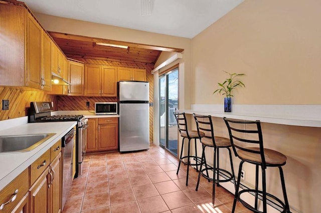 kitchen featuring light tile patterned flooring, under cabinet range hood, stainless steel appliances, light countertops, and brown cabinetry