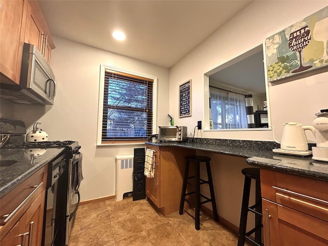 kitchen with baseboards, dark stone counters, radiator, appliances with stainless steel finishes, and brown cabinets