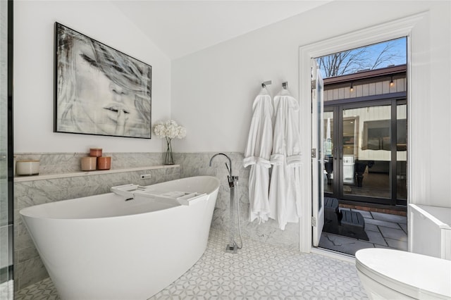 full bath with a wainscoted wall, toilet, tile walls, lofted ceiling, and a soaking tub