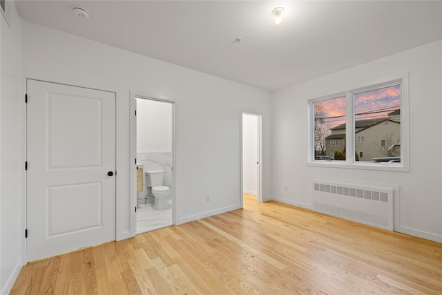 unfurnished bedroom featuring radiator heating unit, light wood-type flooring, ensuite bath, and baseboards