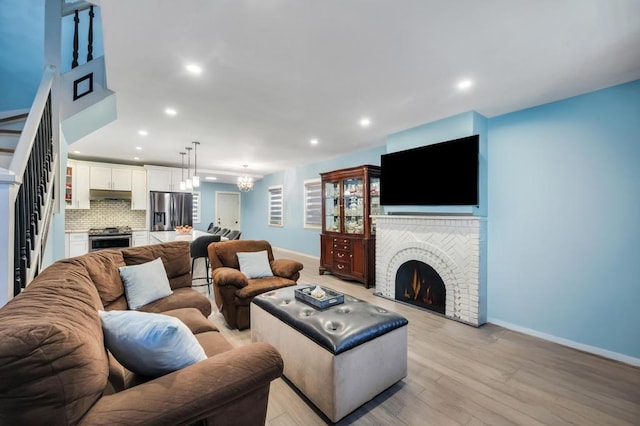 living area with an inviting chandelier, a brick fireplace, light wood-type flooring, baseboards, and stairs
