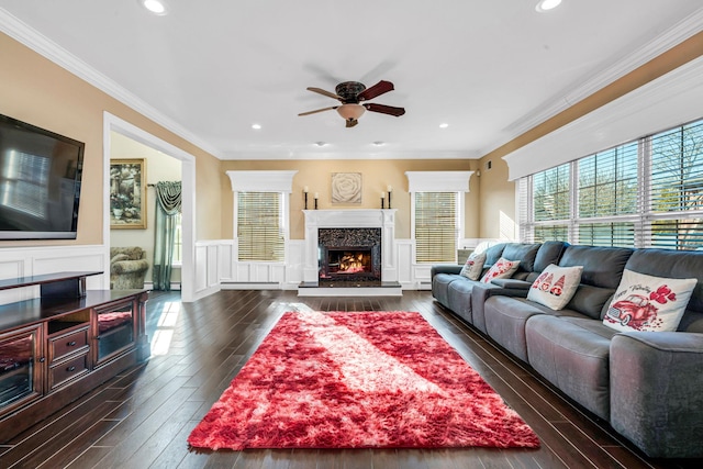 living area with dark wood-style flooring, a wainscoted wall, a premium fireplace, ornamental molding, and a ceiling fan