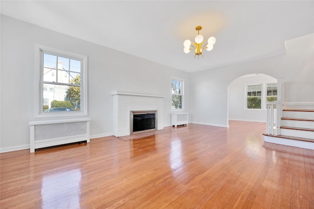 unfurnished living room featuring radiator, an inviting chandelier, light wood-style floors, a brick fireplace, and stairs