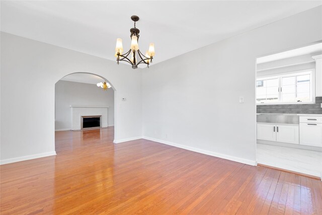 unfurnished living room with arched walkways, light wood-style flooring, baseboards, a brick fireplace, and an inviting chandelier