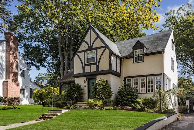 tudor-style house with a shingled roof, a front yard, and stucco siding