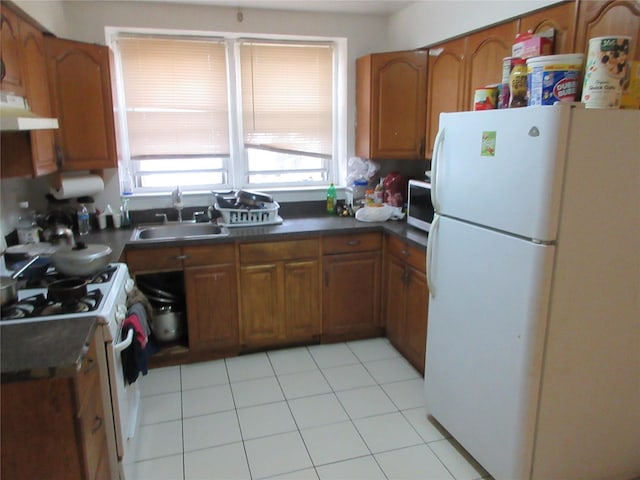 kitchen with white appliances, brown cabinetry, dark countertops, under cabinet range hood, and a sink