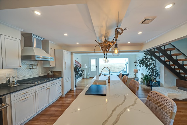 kitchen featuring wood finished floors, stainless steel gas cooktop, crown molding, wall chimney range hood, and tasteful backsplash