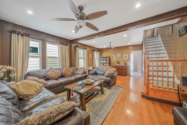 living area with ceiling fan with notable chandelier, wood walls, stairway, light wood finished floors, and beamed ceiling