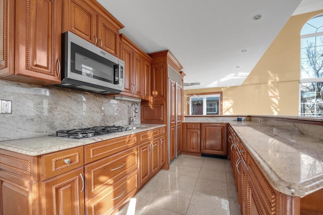 kitchen with light tile patterned floors, stainless steel appliances, brown cabinets, light stone countertops, and tasteful backsplash