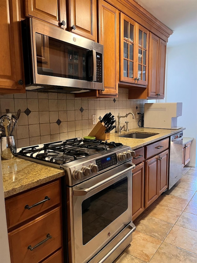 kitchen with decorative backsplash, light stone counters, brown cabinets, stainless steel appliances, and a sink