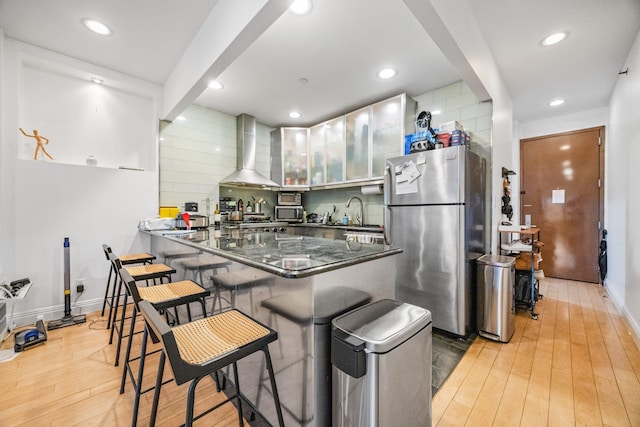 kitchen featuring a peninsula, appliances with stainless steel finishes, decorative backsplash, wall chimney exhaust hood, and light wood finished floors