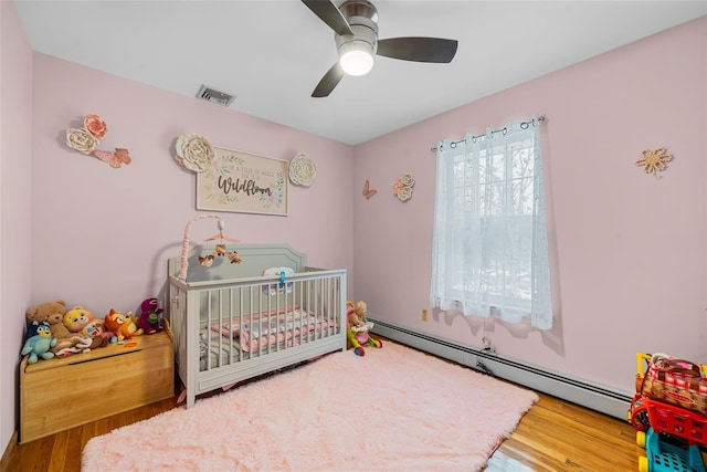 bedroom with wood finished floors, visible vents, a baseboard radiator, ceiling fan, and a nursery area