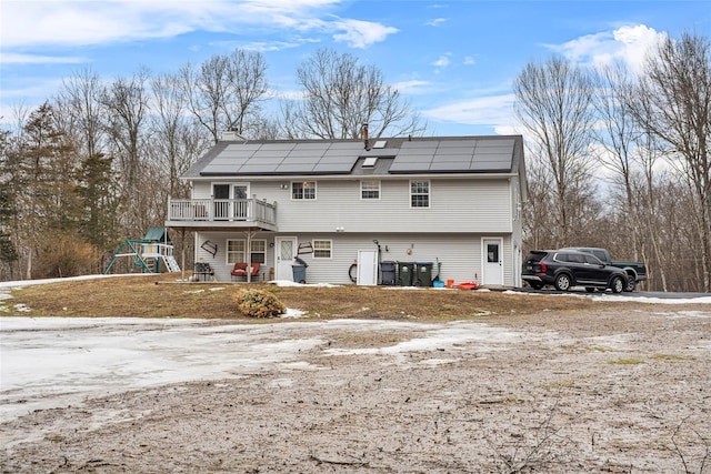 rear view of house featuring a wooden deck, a playground, roof mounted solar panels, and a chimney