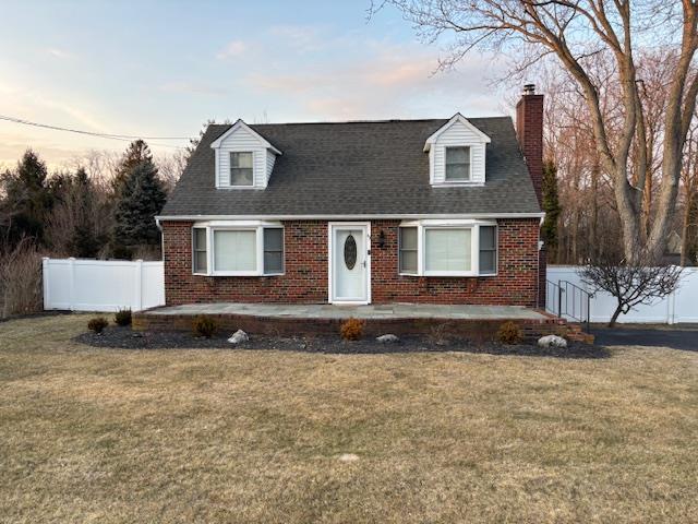 cape cod home featuring brick siding, a chimney, roof with shingles, fence, and a front yard