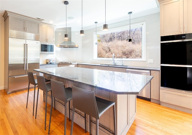 kitchen featuring light wood-style flooring, built in appliances, wall chimney range hood, a kitchen bar, and a sink