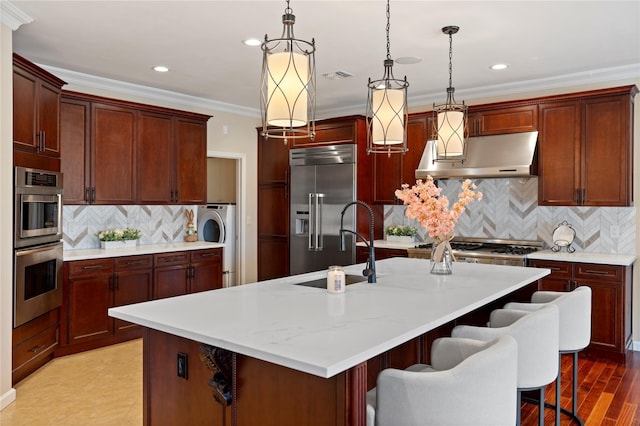 kitchen featuring a kitchen island with sink, under cabinet range hood, stainless steel appliances, washer / clothes dryer, and decorative light fixtures