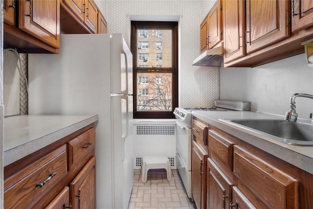 kitchen featuring brown cabinetry, a sink, white appliances, under cabinet range hood, and wallpapered walls