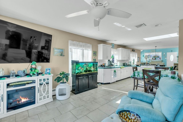 living area featuring a skylight, visible vents, a glass covered fireplace, light tile patterned flooring, and ceiling fan