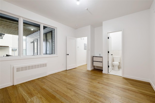 unfurnished bedroom with connected bathroom, baseboards, radiator heating unit, and light wood-style floors