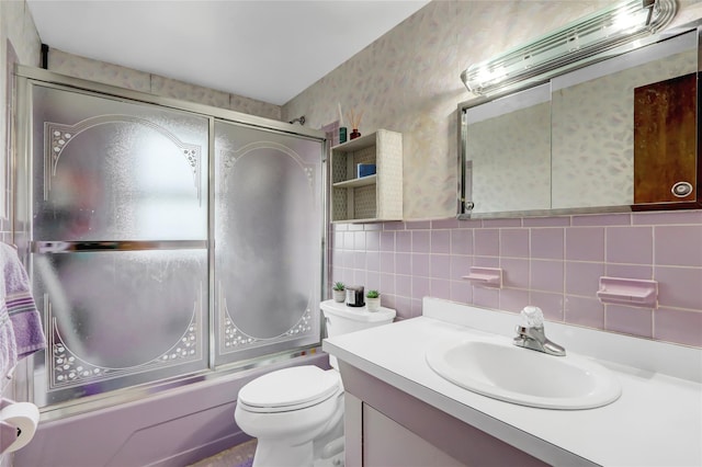 bathroom featuring wallpapered walls, toilet, vanity, shower / bath combination with glass door, and tile walls