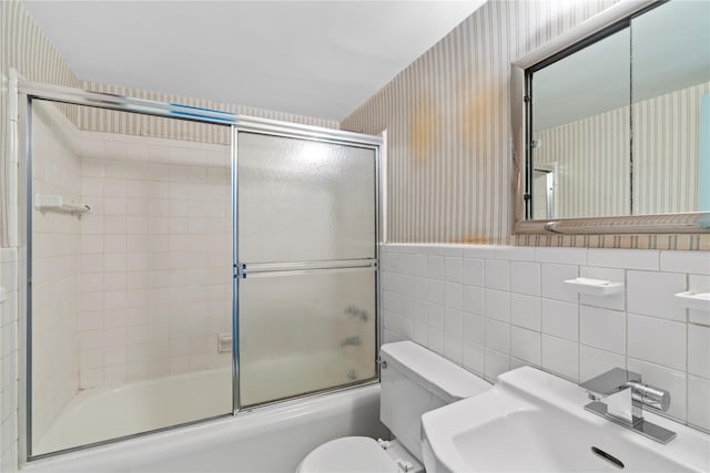 full bathroom with wallpapered walls, bath / shower combo with glass door, a sink, tile walls, and toilet