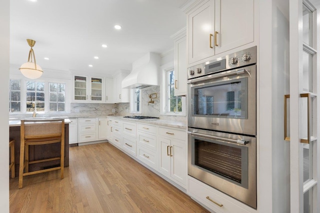 kitchen featuring custom exhaust hood, gas stovetop, decorative backsplash, white cabinets, and double oven