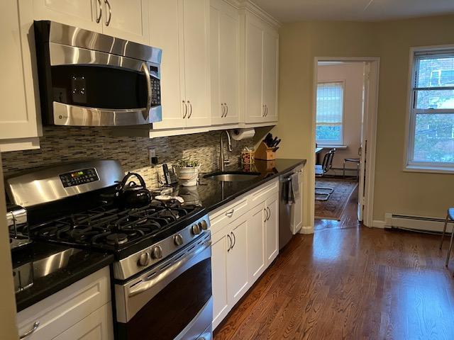 20 Chapel Pl Unit 2K, Great Neck, NY 11021 - kitchen with appliances with stainless steel finishes, a baseboard heating unit, dark wood-type flooring, tasteful backsplash, and white cabinets