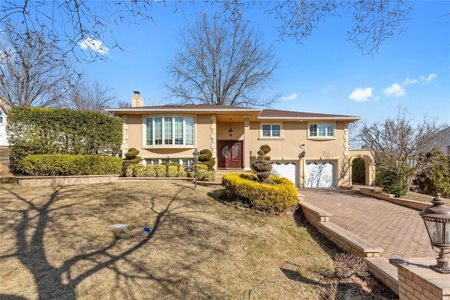 raised ranch featuring driveway, a chimney, an attached garage, and stucco siding