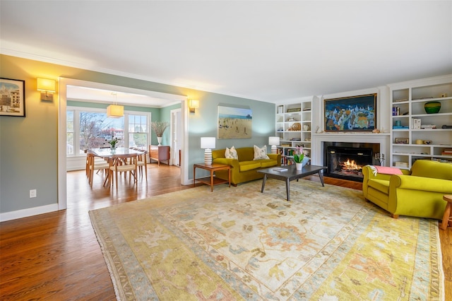 living room with baseboards, a glass covered fireplace, wood finished floors, crown molding, and built in shelves