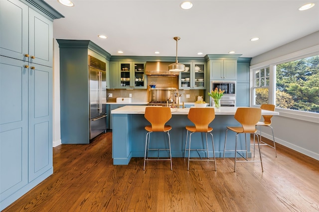 kitchen featuring green cabinetry, wall chimney exhaust hood, built in appliances, light countertops, and backsplash