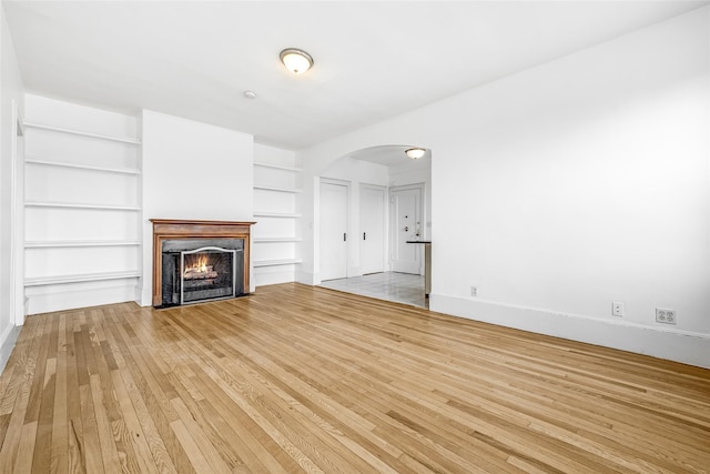 unfurnished living room featuring baseboards, built in features, a lit fireplace, arched walkways, and wood-type flooring