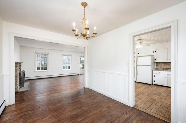 unfurnished dining area with dark wood-style flooring, a wainscoted wall, a baseboard heating unit, a brick fireplace, and ceiling fan with notable chandelier