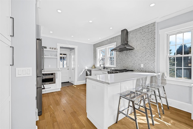 kitchen featuring a peninsula, ornamental molding, stainless steel appliances, light wood-style floors, and wall chimney range hood