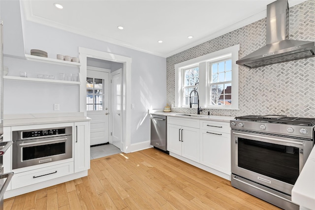 kitchen with crown molding, wall chimney range hood, light wood-style flooring, stainless steel appliances, and a sink