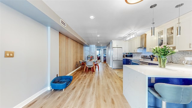 kitchen featuring decorative light fixtures, stainless steel appliances, decorative backsplash, light wood-style floors, and wall chimney range hood