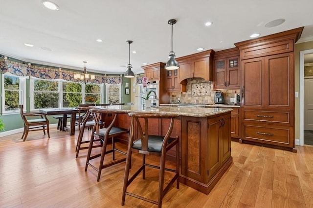 kitchen featuring premium range hood, brown cabinets, light wood-style flooring, and a center island with sink