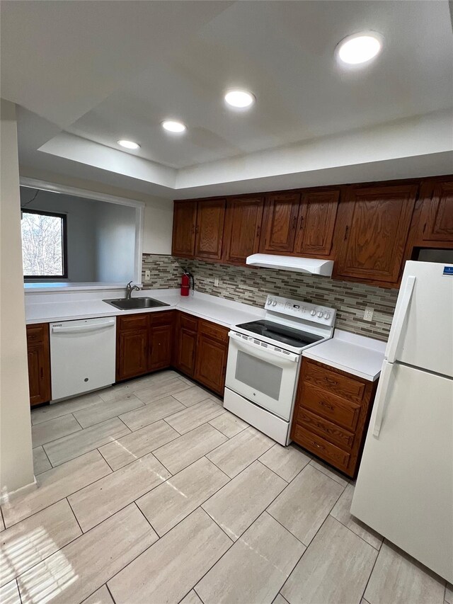 kitchen with under cabinet range hood, decorative backsplash, white appliances, a raised ceiling, and a sink