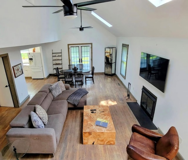 living room featuring high vaulted ceiling, a skylight, a fireplace with flush hearth, wood finished floors, and a ceiling fan