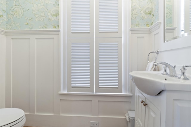 half bath with vanity, toilet, wainscoting, and wallpapered walls