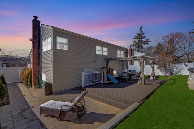 back of house at dusk with a lawn, a fenced backyard, a chimney, a wooden deck, and a pergola