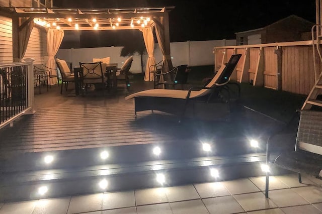 patio at night featuring a fenced backyard, a wooden deck, a pergola, and outdoor dining space