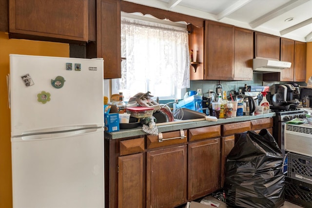 kitchen featuring under cabinet range hood, a sink, freestanding refrigerator, decorative backsplash, and brown cabinetry