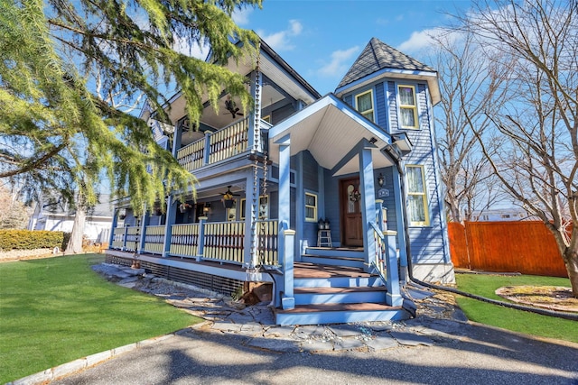 victorian-style house featuring a porch, a front yard, ceiling fan, a balcony, and fence