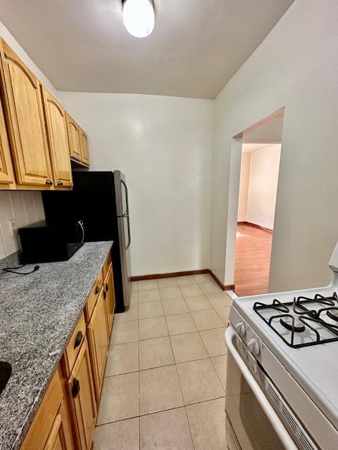 kitchen with light tile patterned floors, white gas stove, baseboards, freestanding refrigerator, and dark stone counters