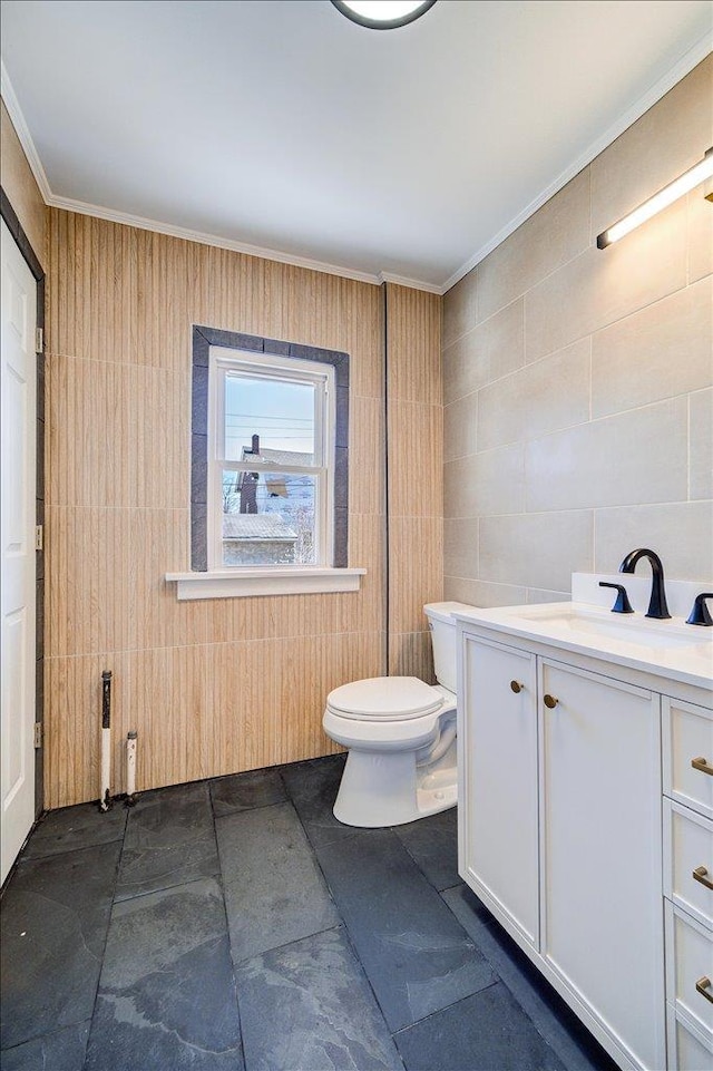 bathroom featuring stone finish flooring, toilet, vanity, ornamental molding, and tile walls