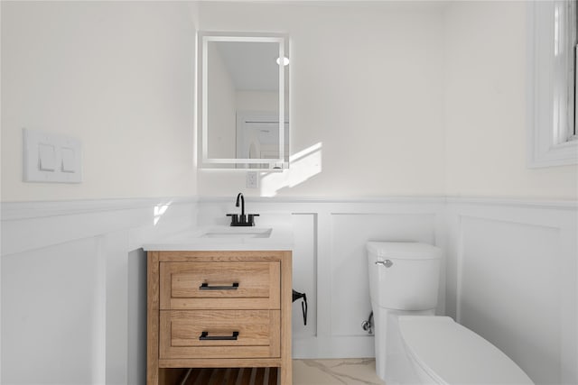 bathroom with wainscoting, toilet, marble finish floor, vanity, and a decorative wall