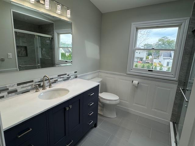 bathroom featuring a stall shower, toilet, a wainscoted wall, vanity, and a decorative wall