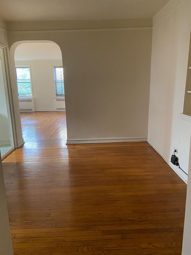 unfurnished room featuring arched walkways, radiator heating unit, wood finished floors, and baseboards